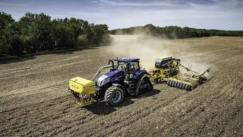 En blå New Holland T8 Genesis traktor trekker landbruksutstyr på en støvete åker.