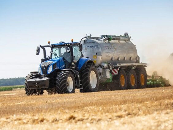 En blå New Holland T8 Genesis traktor trekker en stor Joskin tankvogn på åkeren.
