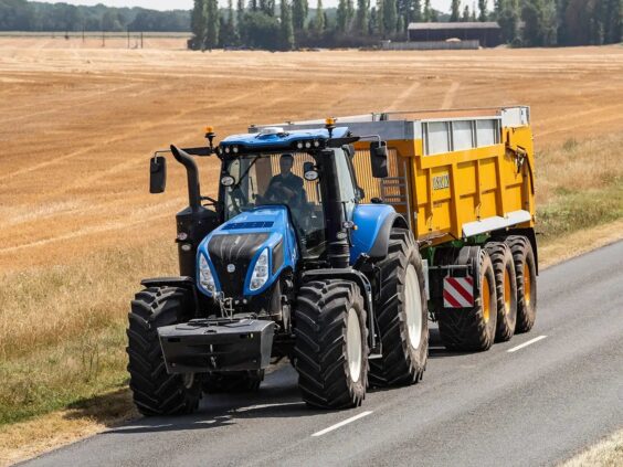 En blå New Holland T8 Genesis traktor som kjører på en asfaltert vei med tilhenger.