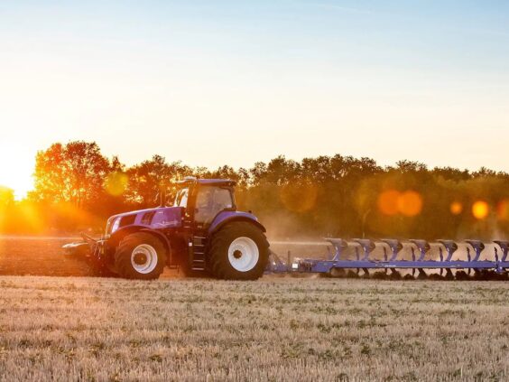 En blå New Holland T8 Genesis traktor pløyer en åker i vakkert solnedgangslys.