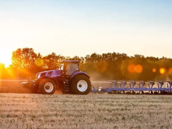 En blå New Holland T8 Genesis traktor pløyer en åker i vakkert solnedgangslys.