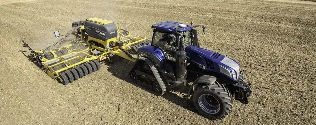 En blå New Holland T8 Genesis traktor trekker en gul Bednar såmaskin på åkeren.