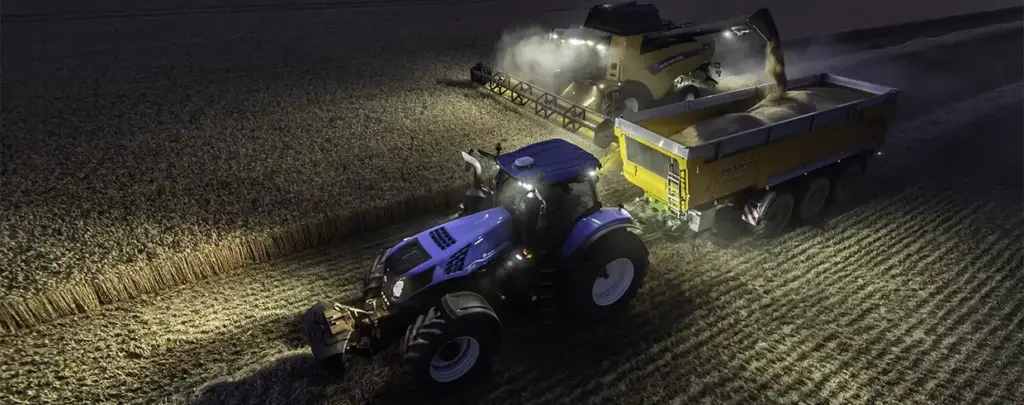 Blå New Holland T8 Genesis traktor som trekker en gul tilhenger under nattlig kornhøsting.