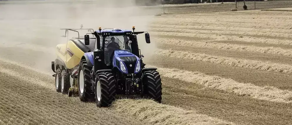 Blå New Holland T8 Genesis traktor trekker en gul halmpresse på en tørr åker.
