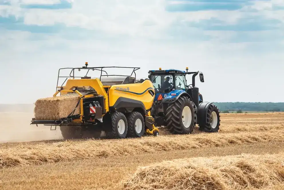 Blå New Holland T8 Genesis traktor med gul ballepresse i arbeid på en åker.
