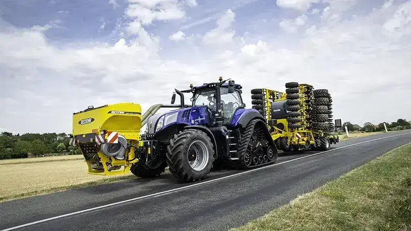 Blå New Holland T8 Genesis traktor med gult landbruksutstyr på en asfaltert vei.
