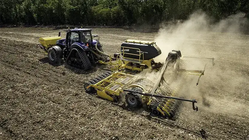 En blå New Holland T8 Genesis traktor trekker en gul såmaskin på åkeren.