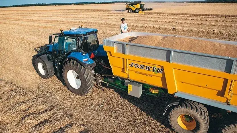 Blå New Holland T8 Genesis traktor med gul Joskin-henger fylt med korn på åkeren.