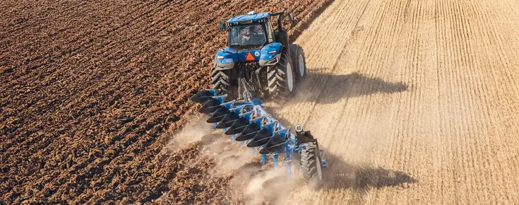 Blå New Holland T8 Genesis traktor som pløyer en stor åker sett ovenfra.