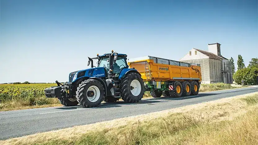 En blå New Holland T8 Genesis traktor med en gul Joskin tilhenger på veien.
