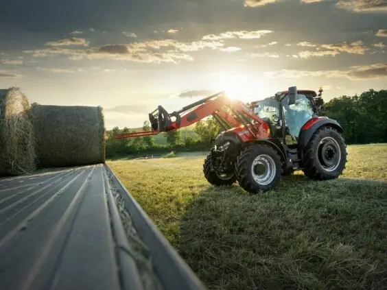 Rød Case IH traktor fra Vestrum-serien laster høyballer på en tilhenger i varm solnedgang.