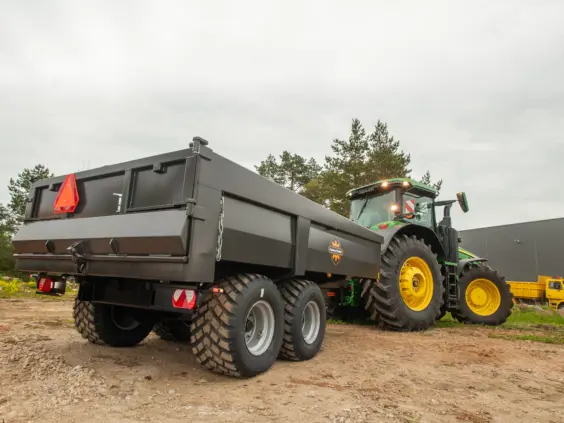 En mørk Palmse PT1300E dumperhenger koblet til en grønn John Deere traktor utendørs.