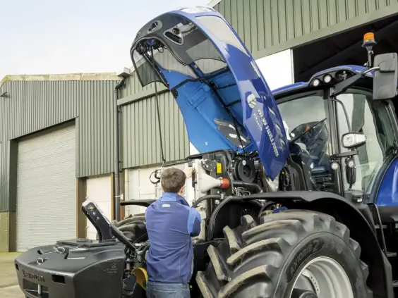 En mekaniker inspiserer motoren på en blå New Holland T7 Heavy Duty traktor utendørs.