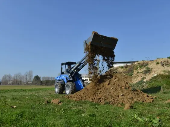 En blå MultiOne 11-serie kompaktlaster tipper jord på en grønn slette.