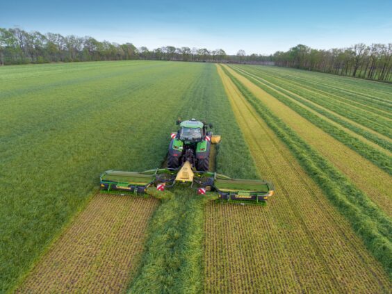En grønn traktor med KRONE EasyCut B slåmaskin klipper gress på et stort jorde.