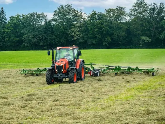 Oransje traktor som vender høy på et grønt jorde med skog i bakgrunnen.