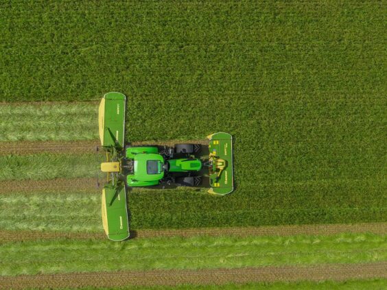 Traktor med KRONE EasyCut B slåmaskin klipper en stor grønn eng sett ovenfra.
