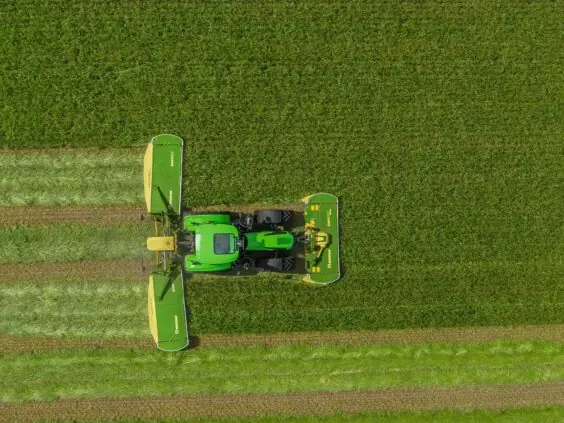 Traktor med KRONE EasyCut B slåmaskin klipper en stor grønn eng sett ovenfra.