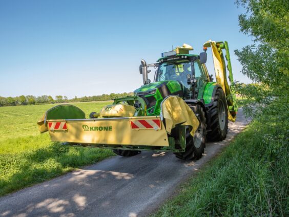 Grønn traktor på en landevei utstyrt med en KRONE EasyCut B slåmaskin.