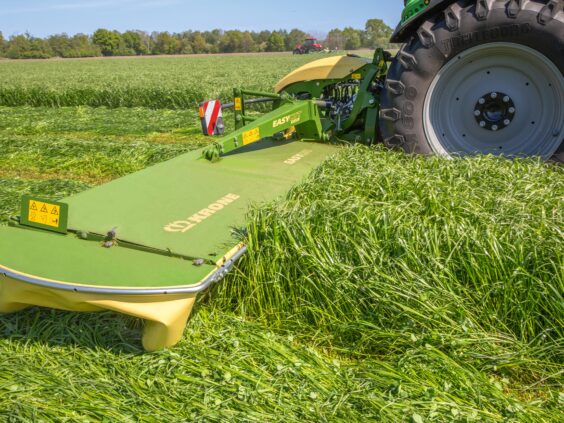 KRONE EasyCut B slåmaskin i aksjon på en grønn åker bak en traktor.