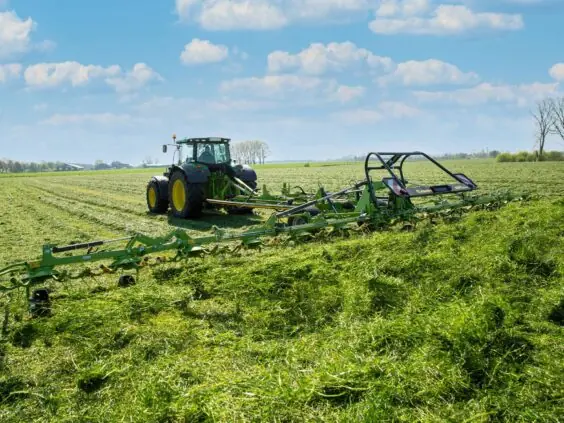 Traktor som trekker en KRONE KWT-serie høyspreder på en grønn eng under blå himmel.