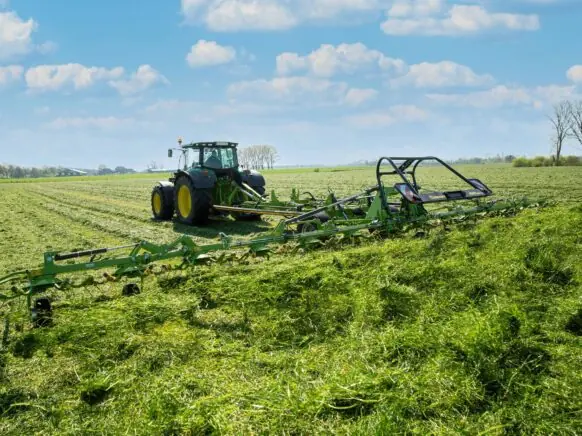 Traktor som trekker en KRONE KWT-serie høyspreder på en grønn eng under blå himmel.