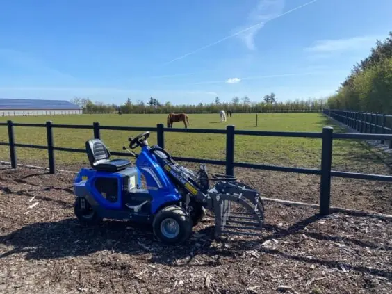 En blå MultiOne 1.1 minilaster parkert ved et gjerde med hester i bakgrunnen.