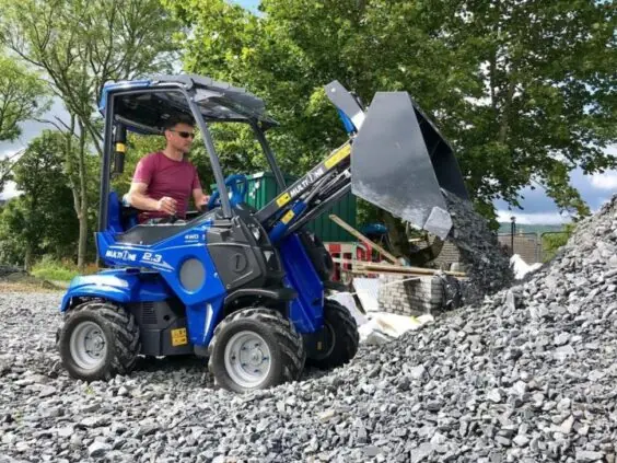 En mann kjører en blå MultiOne 2.3 minilaster og tipper grus på en haug.
