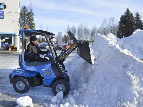 En blå MultiOne 2.3 minilaster rydder en stor snøhaug på en parkeringsplass.