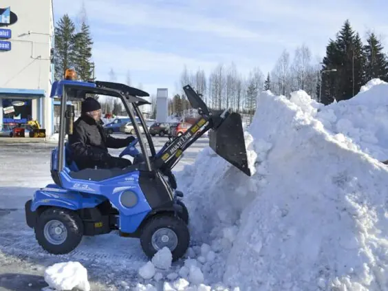 En blå MultiOne 2.3 minilaster rydder en stor snøhaug på en parkeringsplass.