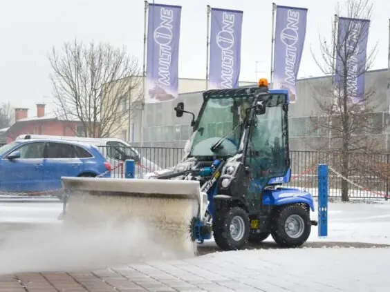 Blå MultiOne 2.3 minilaster med feiebørste som rydder snø på en åpen plass.