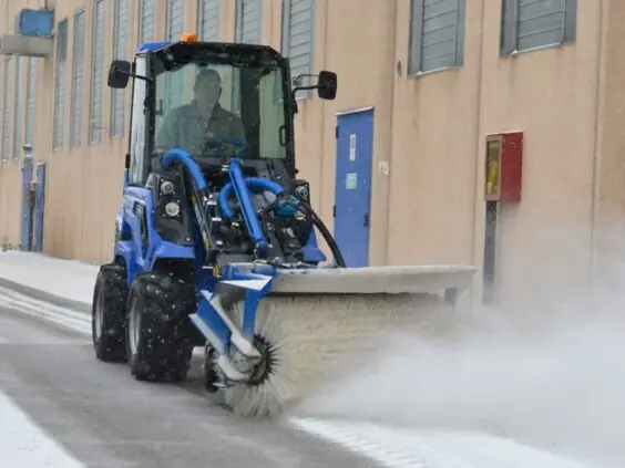Blå MultiOne 6-serie kompaktlaster med feiebørste som rydder snø ved en bygning.