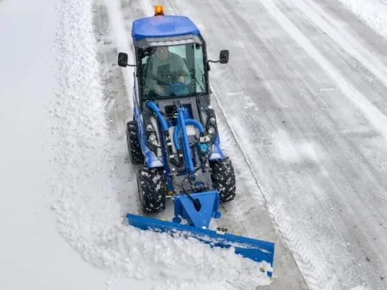 En blå MultiOne 6-serie kompaktlaster rydder snø fra en vei med snøplog.