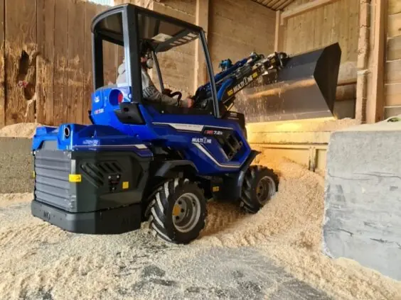 En blå MultiOne 7.2 kompaktlaster flytter sagflis inne i en låve.
