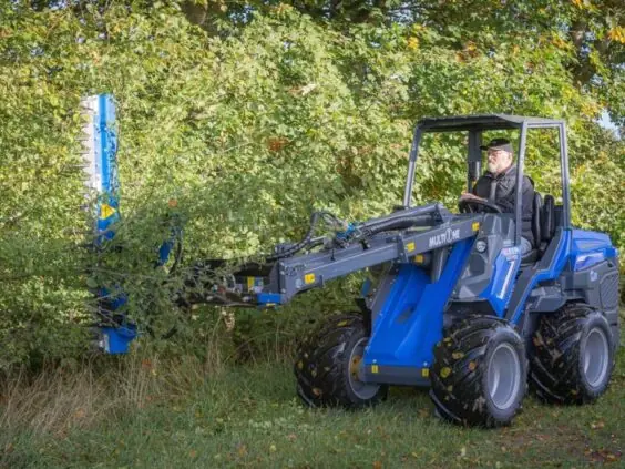 En blå MultiOne 8-serie kompaktlaster klipper en stor hekk med påmontert hekksaks.