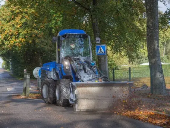 En blå MultiOne 8-serie kompaktlaster feier høstløv fra en asfaltert vei med feiemaskin.