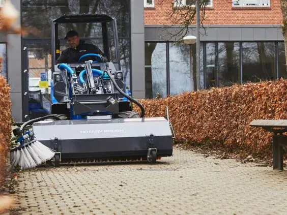 MultiOne EZ-serie elektrisk kompaktlaster utstyrt med feiemaskin på en brolagt gangvei.