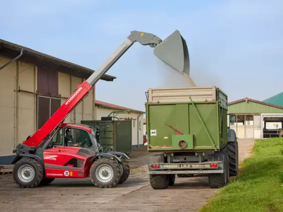 Rød Weidemann teleskoplaster T7035 fyller en grønn landbrukstilhenger med korn på en gård.