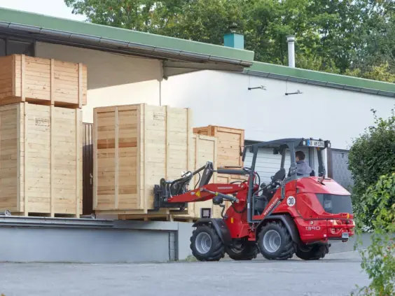 En rød Weidemann Hoftrac 1390 løfter store trekasser ved en lasterampe.