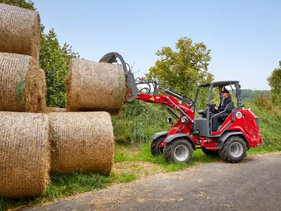 En rød Weidemann Hoftrac 1390 løfter en rundballe for stabling utendørs.
