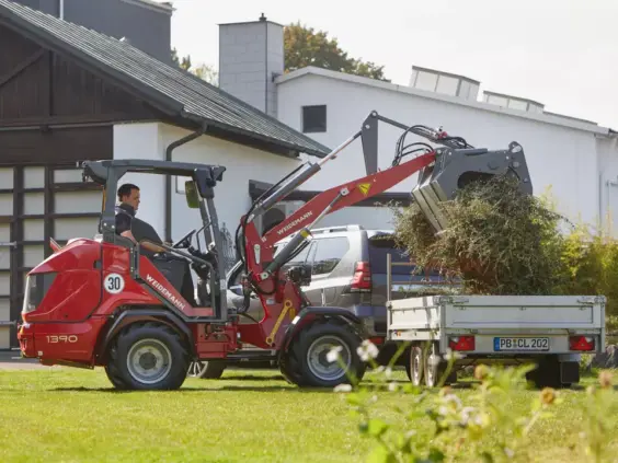 En rød Weidemann Hoftrac 1390 laster hageavfall på en tilhenger i en hage.