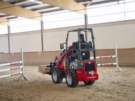 En rød Weidemann Hoftrac 1140 minilaster i en ridehall med ridehindre på sanden.