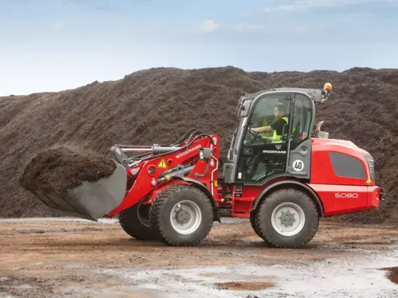 Rød Weidemann Hjullaster 5080 med full skuffe foran en stor jordhaug.