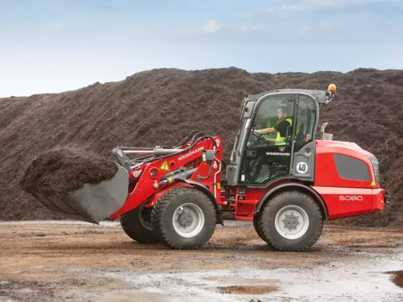 Rød Weidemann Hjullaster 5080 med full skuffe foran en stor jordhaug.