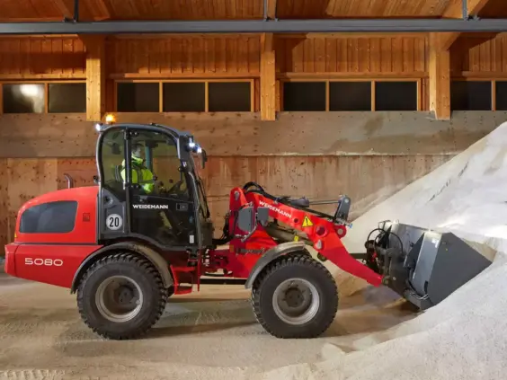 En rød Weidemann Hjullaster 5080 i arbeid med å laste hvitt materiale innendørs.