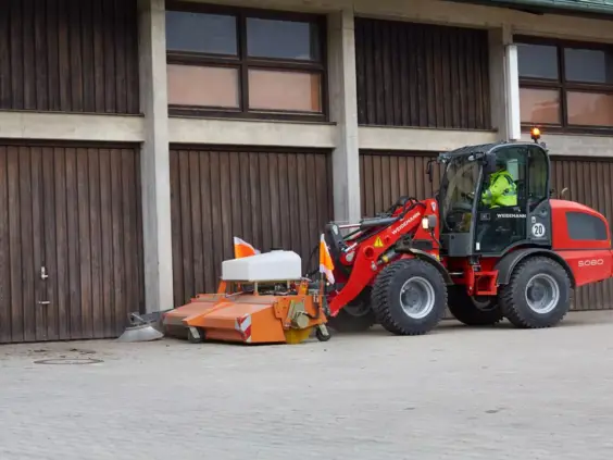 En rød Weidemann Hjullaster 5080 med feieaggregat foran en trebygning.