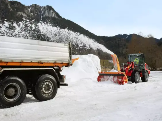 En rød Weidemann Hjullaster 5080 utstyrt med snøfreser som blåser snø på en lastebil.