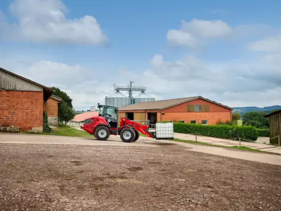En rød Weidemann Hjullaster 4060 frakter en hvit vanntank på en gårdsplass.