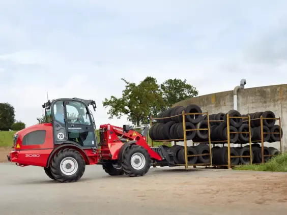 Rød Weidemann Hjullaster 3080 som flytter et stativ med bildekk utendørs.