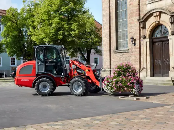 Rød Weidemann Hjullaster 2080 frakter en stor, fargerik blomsterdekorasjon foran en gammel steinbygning.
